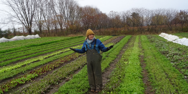 A woman farmer stands in her field, arms outstretched. She is wearing overalls and a hat. At her feet are rows of growing crops.