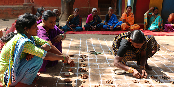 Women crouch outside over a grid drawn with chalk as they place seeds within the squares. The women are wearing colourful saris.