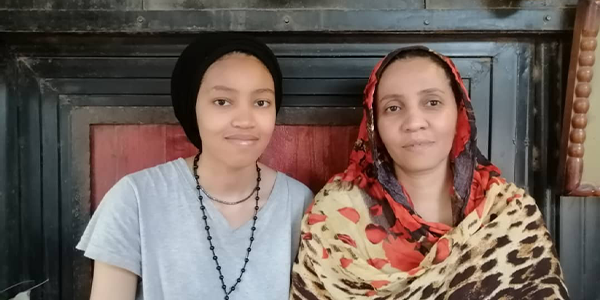 Misson and Jood sit side by side looking at the camera. Misson is wearing a grey t-shirt and black hair wrap. Jood is wearing a colourful wrap.