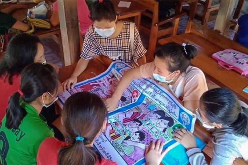 Six jeunes femmes, chacune portant un masque médical, sont assises autour d'une table et regardent un poster représentant deux situations : l'une marquée d'un 