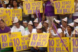 Photo of a large group of people, each holding large yellow signs with purple writing that says "10 YEARS NA ANG RH BILL" and wearing conical cardboard hats.