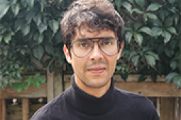 Headshot of a man in glasses and a black turtleneck. Behind him is a wooden fence and a leafy green hedge.
