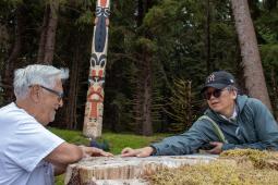 Photo of two people sitting on either side of a wide, moss-covered tree stump. One points at the surface of the stump. In the background, there are tall trees and a totem pole.