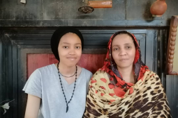 Misson and Jood sit side by side looking at the camera. Misson is wearing a grey t-shirt and black hair wrap. Jood is wearing a colourful wrap.