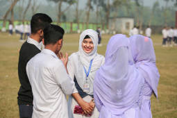 Sanjida Akhter stands in a schoolyard with her hands folded in front of her body, smiling at a group of classmates who stand around her in a circle.