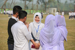 17.	Sanjida Akhter stands in a schoolyard with her hands folded in front of her body, smiling at a group of classmates who stand around her in a circle.