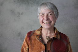 1.	Sari Tudiver smiles brightly at the camera. She is wearing an orange patterned shirt and has short, close-cropped grey hair. Behind her is a grey background. 