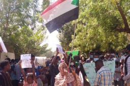 A woman holding a Sudanese flag speaks into a megaphone. She is surrounded by protesters.