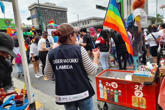 Photo of the back of a person wearing a vest that says "obersvatorio lambda." They are at a Pride parade giving out cool treats.