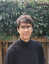 Headshot of a man in glasses and a black turtleneck. Behind him is a wooden fence and a leafy green hedge.