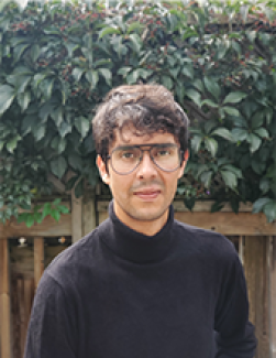 Headshot of a man in glasses and a black turtleneck. Behind him is a wooden fence and a leafy green hedge.